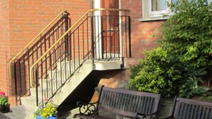 A brick building with a black metal staircase leading to a wooden door. There is a bench and a potted plant with yellow flowers on the ground. - Town House, situated in the village Salthill close to the seafront. (Galway)
