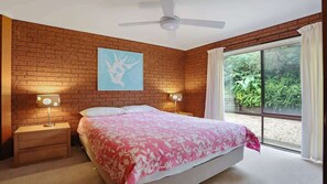 Ein Schlafzimmer mit einer Ziegelwand, einem Bett mit blumigem Überwurf, zwei Nachttischlampen, einem Deckenventilator und einem großen Fenster mit Vorhängen.
