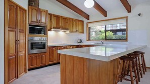 A kitchen with wooden cabinets, a built-in oven, a microwave, a large island with stools, and a window with a view of greenery.