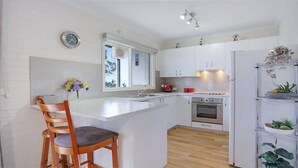 A modern kitchen with white cabinets, a wooden chair, a built-in oven, and a refrigerator. - North Terrace 9 (Merimbula)