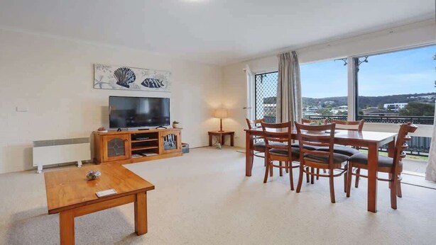 A living room with a TV, wooden furniture, a table, and a balcony with a view. - North Terrace 9 (Merimbula)