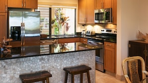 A kitchen with wooden cabinets, stainless steel appliances, a large island with stools, and a stone backsplash. - Stunning One Bedroom in the Heart of Wailea Ekahi Village Unit 28B (Kihei)