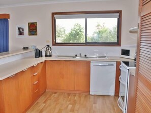A compact kitchen with wooden cabinets, a white countertop, a window with blinds, a sink, a microwave, and a dishwasher.