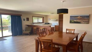 Een eetgedeelte met een houten tafel en stoelen, een keuken met een magnetron, en een groot raam met gordijnen.