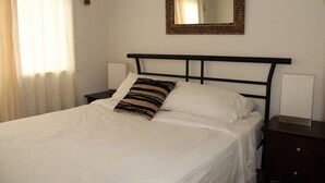Ein ordentlich bezogenes Bett mit weißer Bettwäsche und einem gestreiften Kopfkissen, ein Nachttisch mit Lampe und ein Spiegel an der Wand.