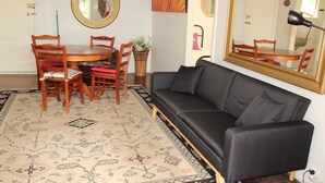 Ein Wohnzimmer mit einer schwarzen Couch, einem Holzespeisetisch mit Stühlen, einem gemusterten Teppich und einem großen Spiegel an der Wand.
