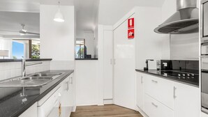 A modern kitchen with white cabinets, a black countertop, stainless steel appliances, and a ceiling fan. - Byron Bay Accom - The Palms 4 - 54 Lawson Street (Byron Shire)