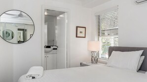 A bedroom with a bed, a bedside table with a lamp, a window with blinds, a door leading to a bathroom, and a round mirror on the wall. - Byron Bay Accom - The Palms 4 - 54 Lawson Street (Byron Shire)