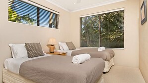 Quarto de hotel com duas camas, uma mesa de cabeceira, abajur e janelas amplas com persianas.