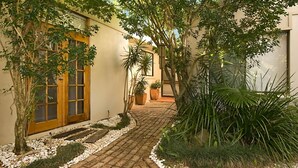 Uma trilha pavimentada leva a um edifício com porta e janelas de madeira, cercado por vegetação exuberante e plantas em vasos.