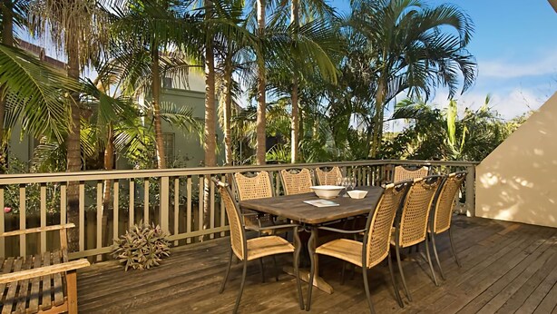 Um deck externo com mesa de madeira e cadeiras de vime, cercado por palmeiras e um muro branco.