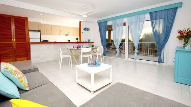 Una sala de estar moderna con sofá, una mesa de comedor con sillas, zona de cocina y un balcón con cortinas azules.
