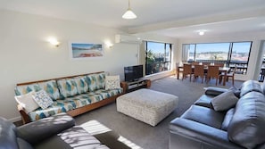 A living room with a sofa, ottoman, TV, and dining area with chairs and a table. - The Escape (Merimbula)