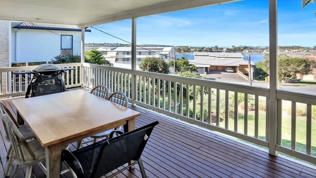 A covered balcony with a table and chairs, overlooking a residential area with houses and a body of water. - The Escape (Merimbula)
