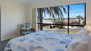 A bedroom with a large window, a bed with a patterned bedspread, a chair, and a view of palm trees and a street. - The Escape (Merimbula)