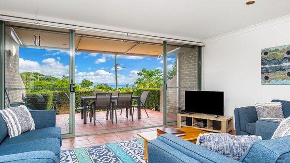 A modern living room with a TV, sofa, and dining area with a view of greenery and a clear sky.