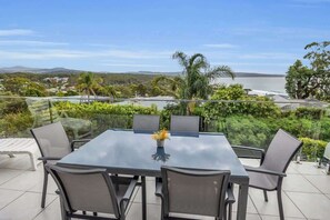 A dining area with a glass table and chairs, overlooking a scenic view of a body of water and hills. - Seaview Pambula Beach (Pambula Beach)