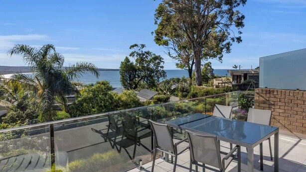 A balcony with a glass table and chairs, overlooking a coastal area with trees and a view of the sea. - Seaview Pambula Beach (Pambula Beach)