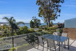 A balcony with a glass table and chairs, overlooking a coastal area with trees and a view of the sea. - Seaview Pambula Beach (Pambula Beach)
