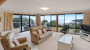 A spacious living room with a large window, a sofa, a television, and a table. - Sullivan's on Kanangra (Pambula Beach)