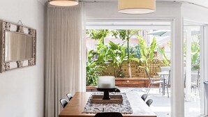 A dining area with a wooden table, chairs, and a cake on a stand. There is a curtain on the left and a view of a garden with plants and a brick wall. - Byron Bay Accom - Eastpoint 2 - 18 - 20 Paterson Lane (Byron Shire)