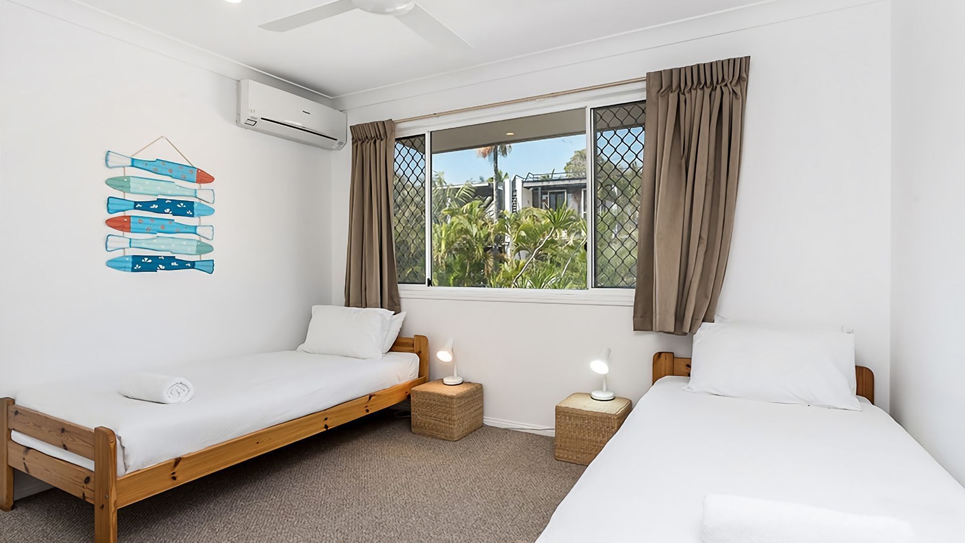 A bedroom with two single beds, a window with curtains, an air conditioning unit, and a wall decoration of boats.