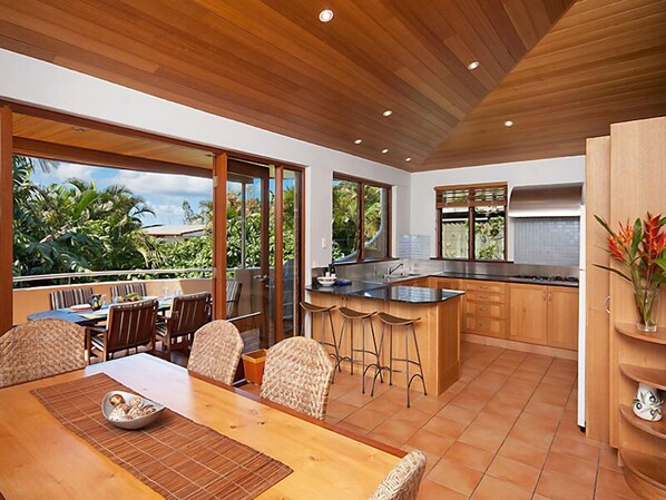 Enjoy meals in the spacious kitchen and dining area with a large island and bar stools, featuring a stainless steel sink, range hood, and wooden cabinets, and a dining table with wicker chairs.