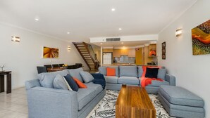 Un saló modern amb un sofà seccional gris, una taula de centre de fusta i una escala que condueix a una cuina.