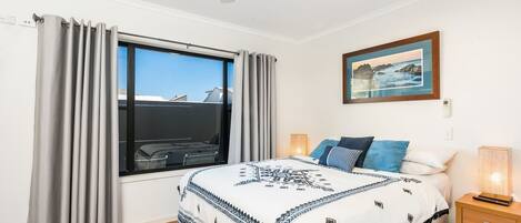 Een slaapkamer met een groot raam, een bed met beddengoed met een patroon, een nachtkastje met een lamp en een ingelijst schilderij aan de muur.