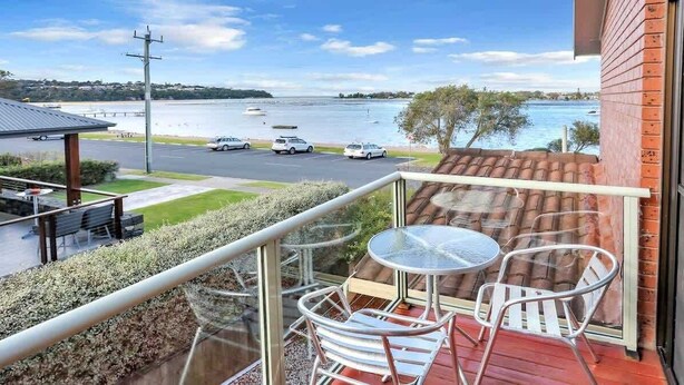 A balcony with a glass railing, a table, and two chairs overlooking a body of water with boats and a parking area.