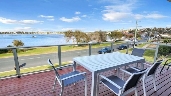 A deck with a table and chairs overlooking a body of water and a road.