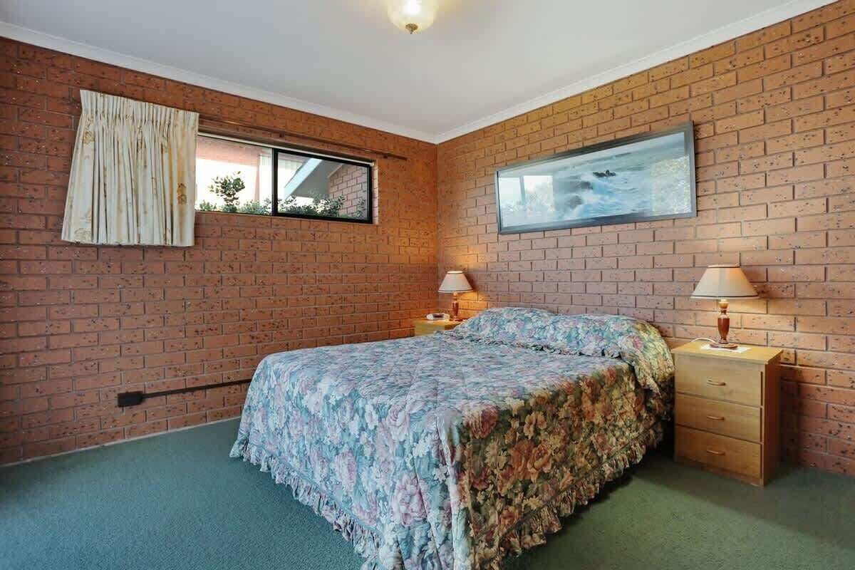 A bedroom with a brick wall, a bed with floral bedding, two bedside tables with lamps, and a window with curtains.