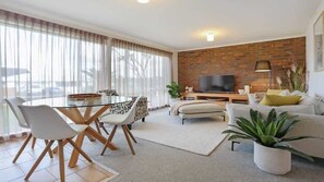 A modern living room with a brick wall, a flat-screen TV, a dining table with chairs, a sofa, and a potted plant.