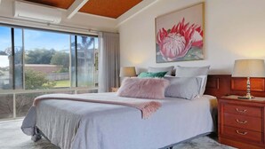 A bedroom with a large bed, a nightstand, a window with a view of the outdoors, and a painting on the wall. - PiPis on Collins (Merimbula)