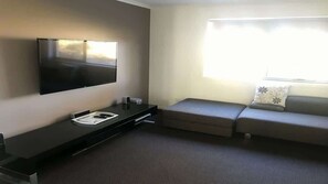 A modern hotel room with a flat-screen TV mounted on the wall, a small desk with a telephone, a sofa, and a window with blinds. - Turaroo (Tura Beach)