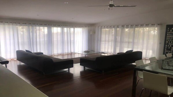 A modern living room with a glass coffee table, two sofas, a dining table with chairs, and large windows with sheer curtains.