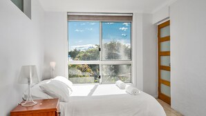 A bedroom with a large window, a bed, a bedside table with a lamp, and a wooden door.