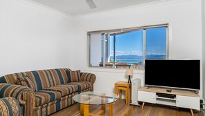 A living room with a sofa, a glass coffee table, a TV on a wooden stand, and a view of the sea through a large window.