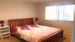 A bedroom with a wooden bed, a nightstand, a lamp, a window with blinds, and a view of the outside. - Coastal Escape (Tura Beach)