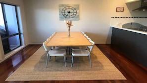 A dining area with a wooden table, white chairs, a clock on the wall, and a kitchen in the background. - Coastal Escape (Tura Beach)