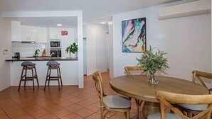 A modern kitchen with white cabinets, a central island, and a dining area with a round wooden table and four wooden chairs. - Byron Bay Accom - Nautilus 1 - 46 Lawson Street (Byron Shire)