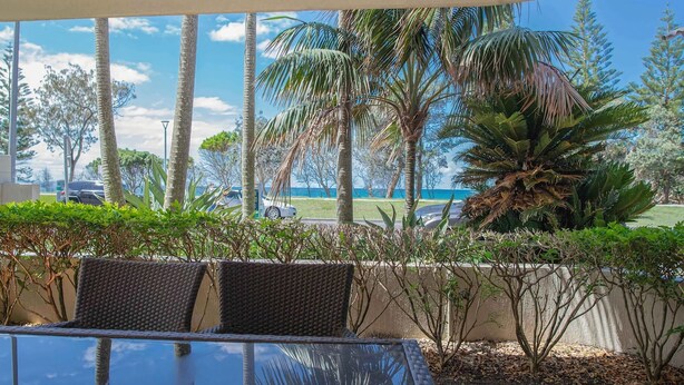 An outdoor seating area with wicker chairs and a glass table, overlooking a parking lot and a view of the ocean. - Byron Bay Accom - Nautilus 1 - 46 Lawson Street (Byron Shire)