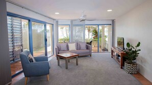 A spacious living room with a grey sofa, a blue armchair, a wooden coffee table, and a TV stand with a plant. - Byron Bay Accom - Nautilus 1 - 46 Lawson Street (Byron Shire)