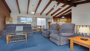 Ein Wohnzimmer mit einer Couch, Sesseln, einem Couchtisch und einer Lampe. Im Hintergrund steht ein Esstisch mit Stühlen.