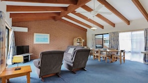 A spacious living area with a brick wall, wooden beams, a television, a sofa, a dining table with chairs, and a bookshelf. - SeaLeka 5 (Merimbula)