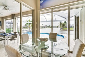 Mesa de jantar com tampo de vidro, cadeiras, uma tigela e vista para uma piscina e palmeiras.
