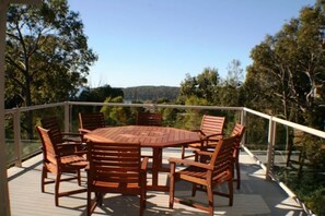 A wooden deck with a round table and chairs, surrounded by glass railings, overlooking a forested area. - Deja Blue - Pambula Beach (Pambula Beach)