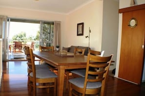 A dining area with a wooden table and chairs, a sofa, and a door with a round mirror. - Deja Blue - Pambula Beach (Pambula Beach)