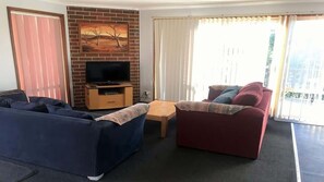 A living room with a sofa, a red couch, a TV on a wooden stand, and a window with curtains.