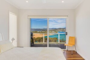 A bedroom with a large bed, a chair, and a sliding glass door leading to a balcony with a view of a beach and mountains.